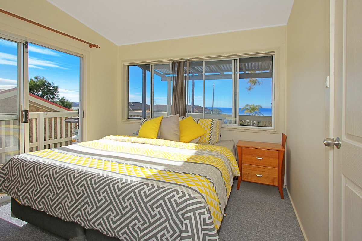 A well-lit bedroom features a spacious bed dressed in a patterned blanket, accented by bright yellow cushions. Large windows provide views of the ocean and allow natural light to fill the room. A wooden nightstand is positioned beside the bed, enhancing the cozy setting.