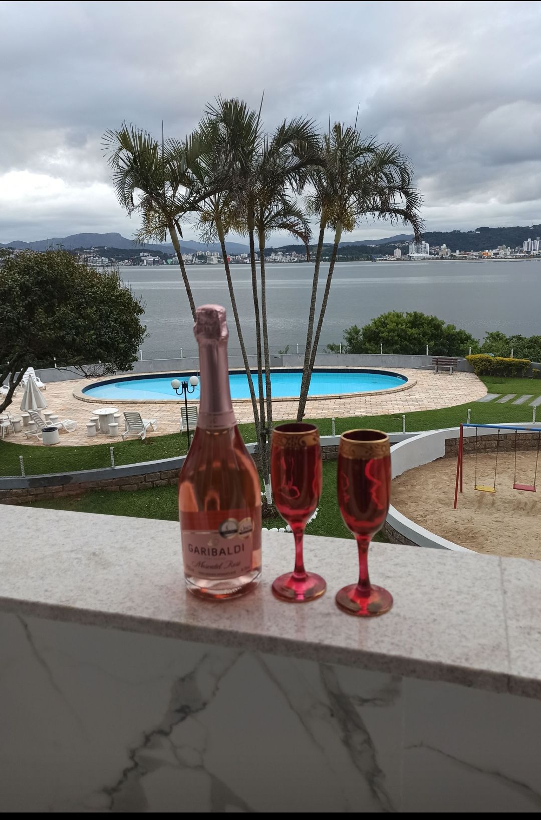 A marble countertop displays a bottle of sparkling wine alongside two elegant glasses. In the background, a sparkling pool is framed by palm trees and offers a panoramic view of the water and distant cityscape.