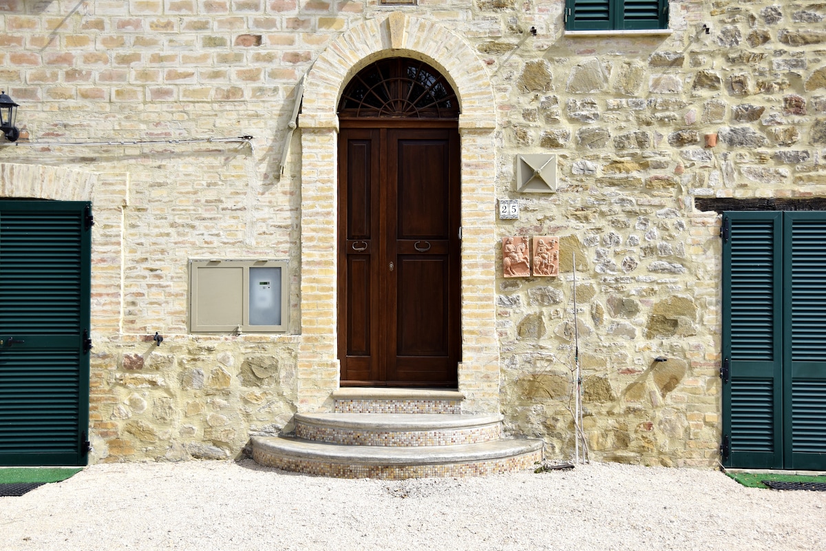 A sturdy wooden door stands as the entrance to a stone-clad building. The door is framed by an arched window and is flanked by two green shutters. A set of stone steps leads to the entrance, with gravel surrounding the base.