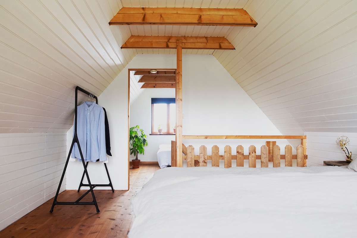 The bright bedroom features a spacious bed with a light-colored duvet. A wooden beam design and a soft, sloped ceiling create an airy feel. A clothing rack showcases a neatly hung shirt, while a small plant adds a touch of greenery. A door leads to another room.