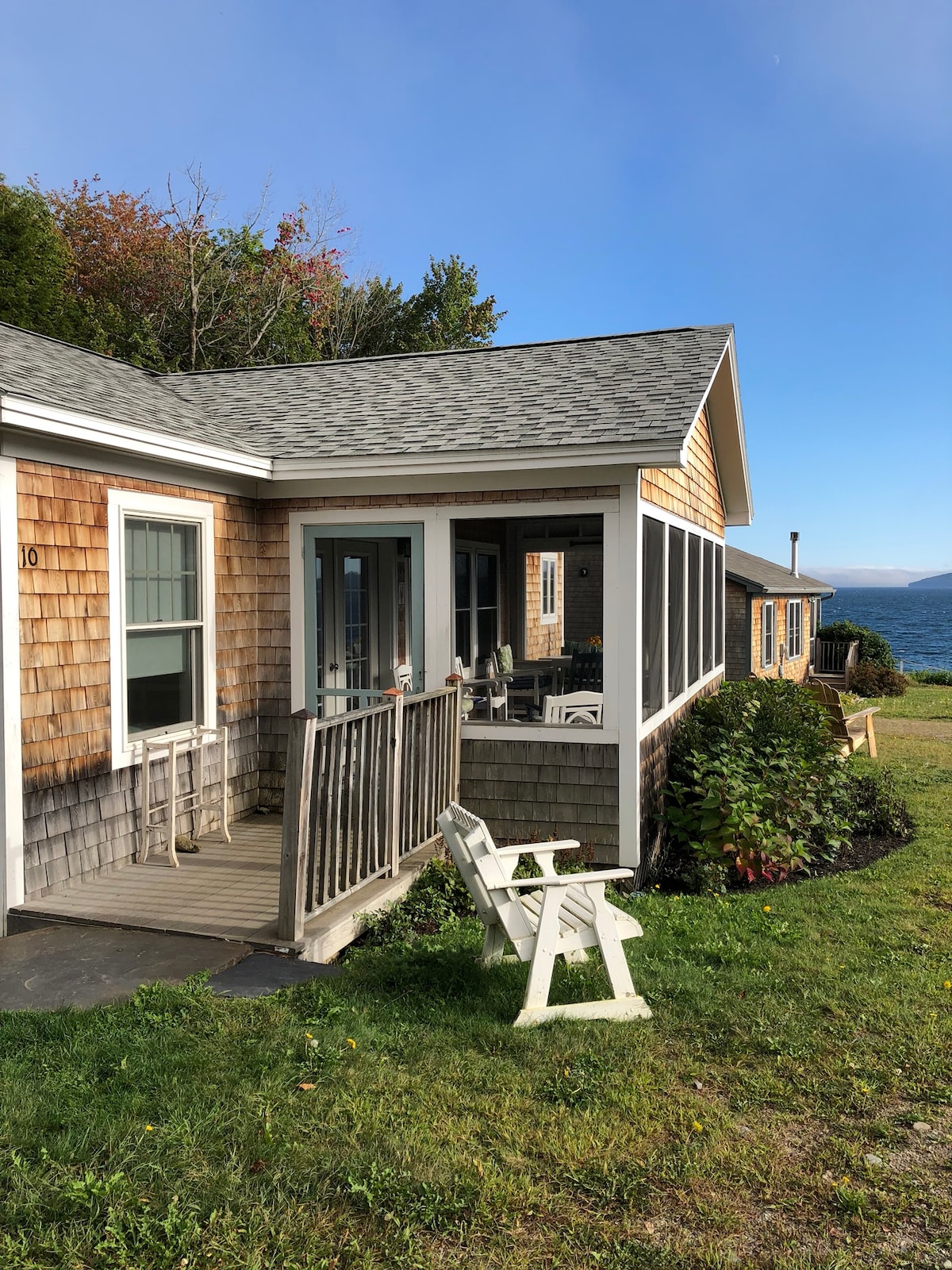 The exterior of the cottage showcases a shingle-style façade with a wooden porch. A white Adirondack chair is positioned on the lawn, overlooking expansive views of Penobscot Bay. The scene is framed by greenery and a clear blue sky, evoking a serene coastal atmosphere.