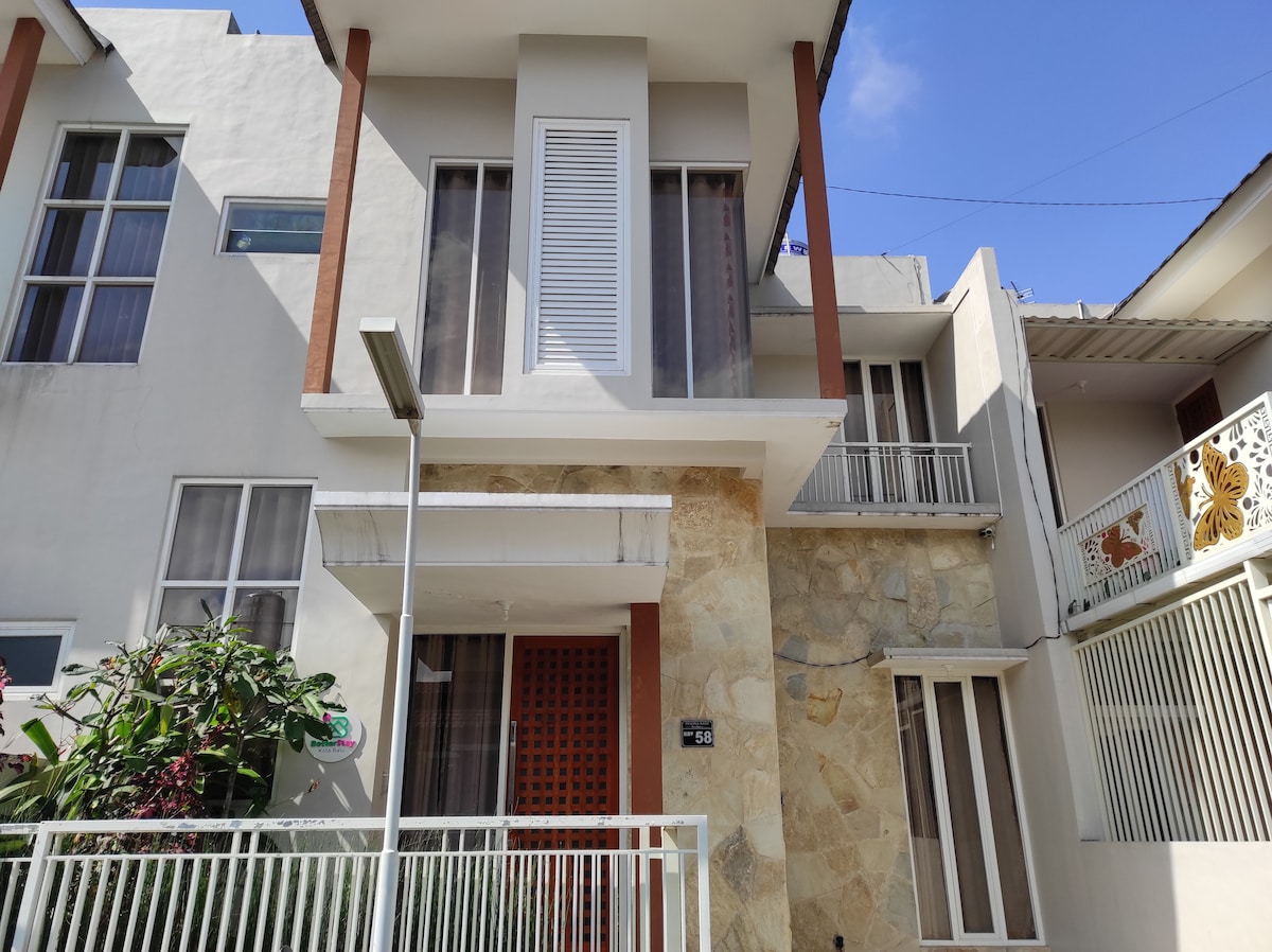 The exterior of the house is showcased, featuring a combination of stone and smooth wall finishes. A wooden door with decorative detailing is framed by large windows on both levels. Lush green plants border the front yard, and the clear blue sky is visible above.