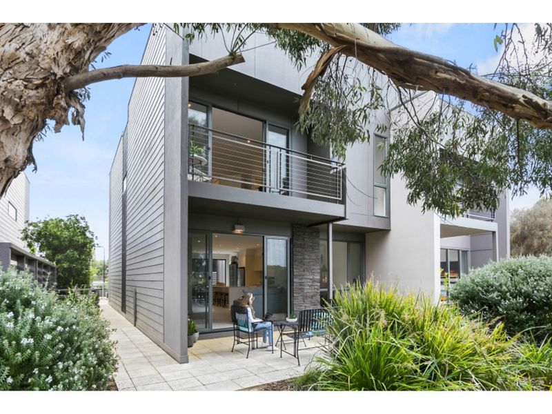 The exterior of a modern two-story condo is shown, featuring a balcony with seating for two. Lush greenery and low-maintenance landscaping enhance the entrance, while large glass doors provide a glimpse into the bright interior space.