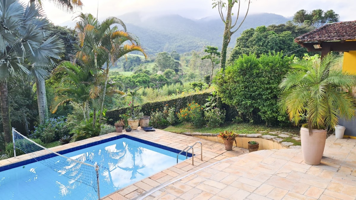 A serene pool is surrounded by natural greenery, showcasing lush palm trees and diverse plants. The landscape includes rolling hills and misty mountains in the background. The pool area features a stone patio and a net for recreational activities.