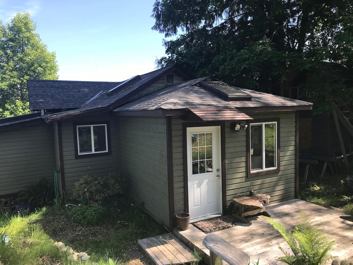 The exterior of the cottage is shown, featuring a welcoming front door framed by large windows. A wooden deck leads to the entrance, surrounded by a garden area with greenery. The roof displays a combination of textures, with trees providing additional shade.