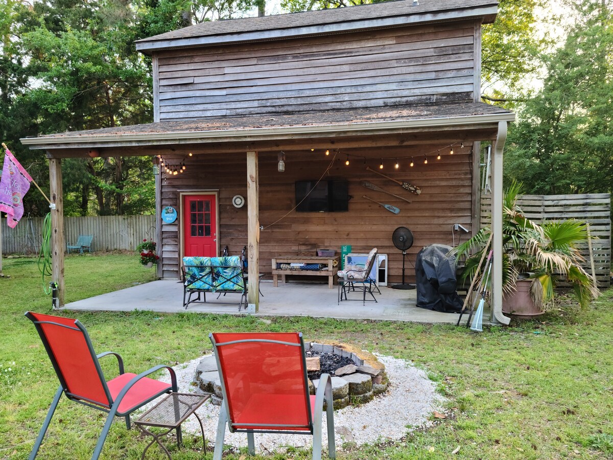 An outdoor living area is featured under a covered porch with wooden walls. Two red chairs are positioned around a fire pit, surrounded by gravel. A TV is mounted on the wall, with string lights illuminating the space. Lush greenery surrounds the area, enhancing a natural setting.