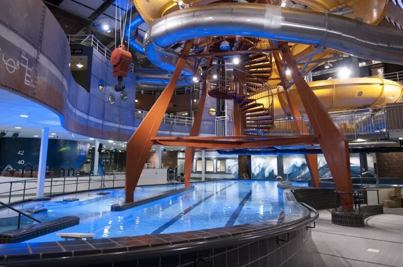 An indoor water park area features a large swimming pool with clear blue water, surrounded by a winding water slide structure and spiral staircase. Industrial-style lighting illuminates the space, creating an energetic atmosphere. Seating areas are present alongside the pool for relaxation.