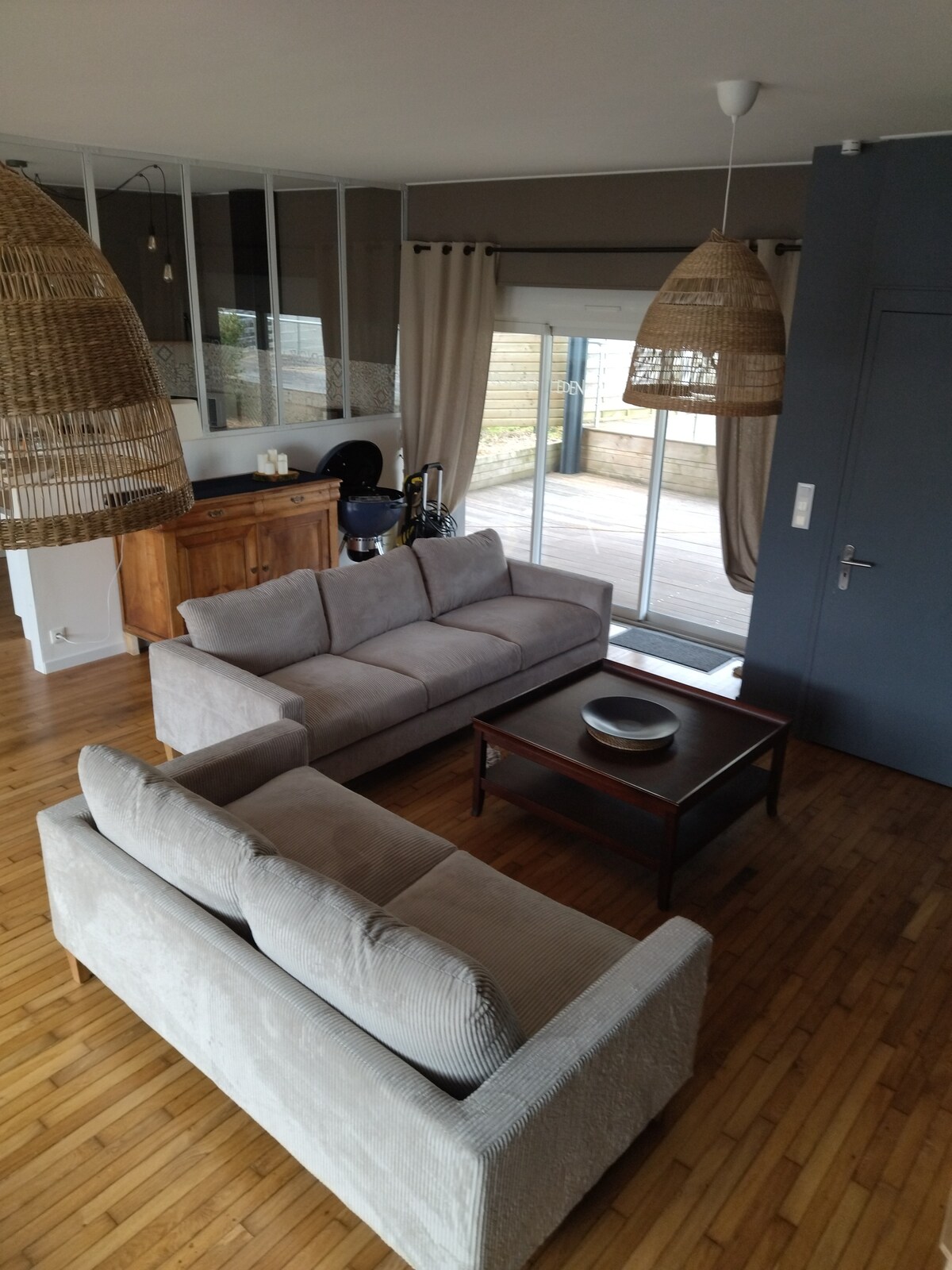 A spacious living area features two light-colored sofas positioned around a wooden coffee table. Large sliding doors provide access to an outdoor deck, allowing natural light to fill the space. Hangings made of woven materials add texture to the room.