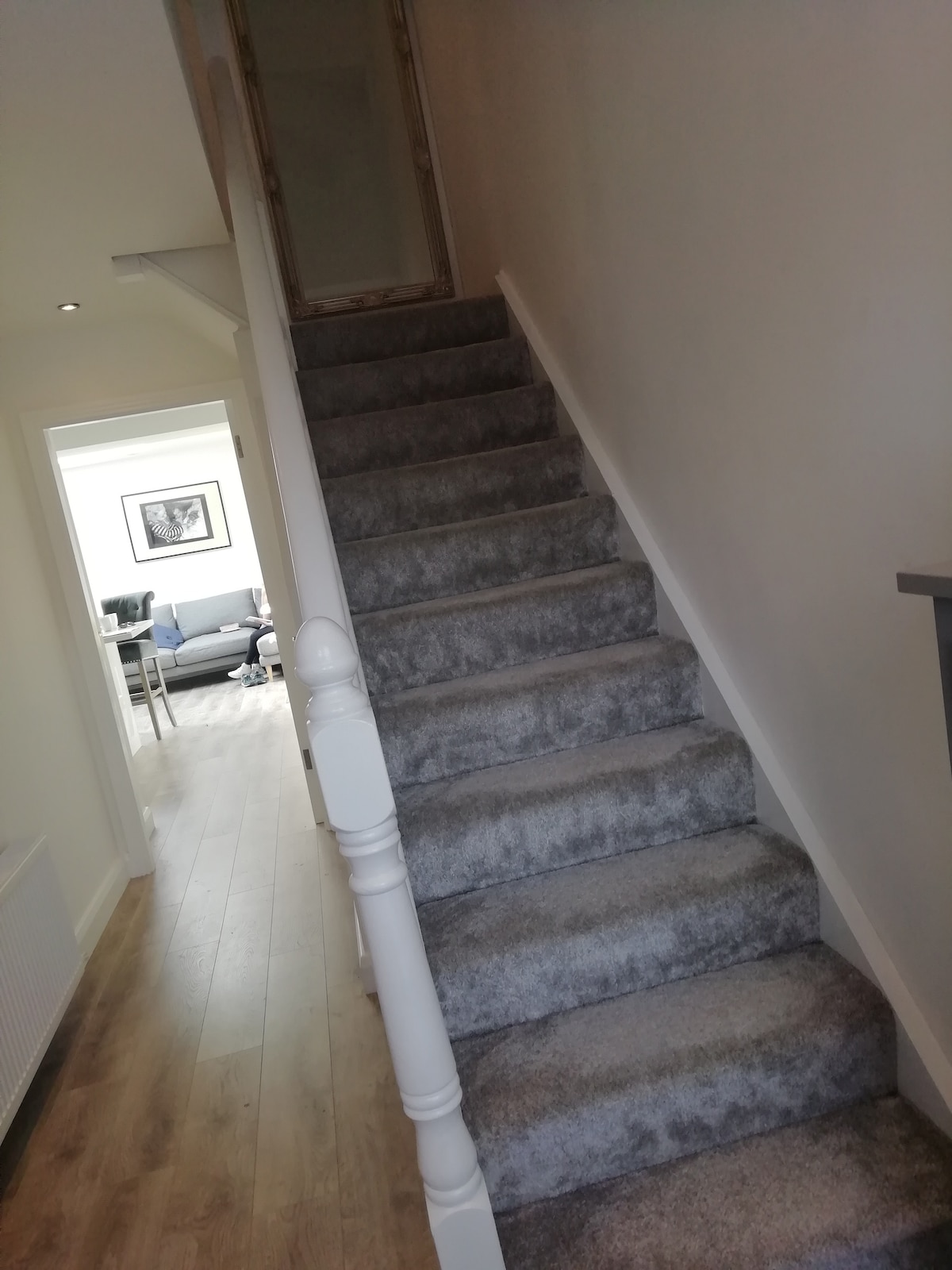 A carpeted staircase leads upward, featuring softly curved banisters. The steps are illuminated by ceiling lights. A glimpse of a living area is visible at the top, showcasing a comfortable seating arrangement and a large window allowing natural light to fill the space.