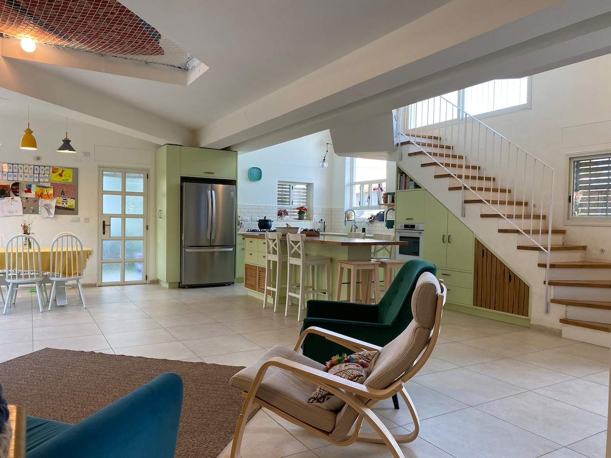 A spacious central living area features an open layout with a large kitchen and dining space. Light-colored furniture and cabinets create a bright atmosphere. A staircase leads to the upper floor, while large windows allow natural light to illuminate the room.
