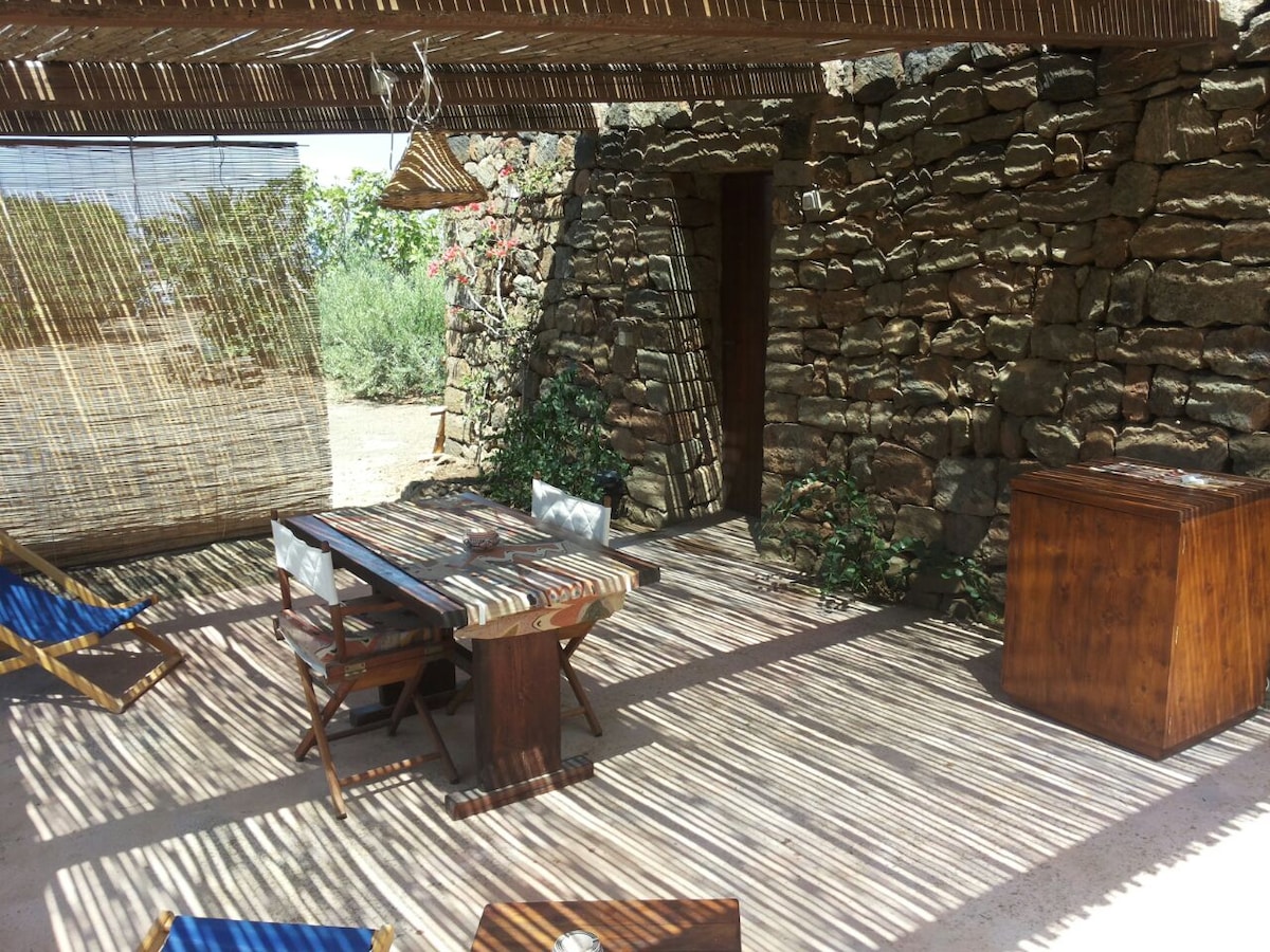 An outdoor terrace is featured, framed by natural stone walls and shaded with a bamboo roof. A wooden dining table accompanied by folding chairs is shown, with greenery and hanging decorations nearby. The flooring is composed of textured light-colored materials.