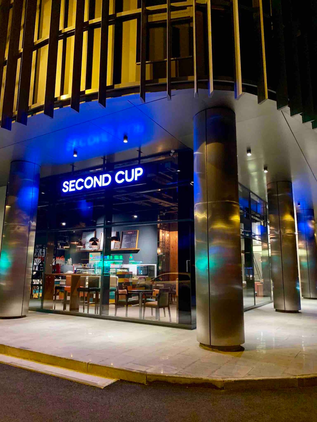 A modern café facade is highlighted by large glass windows and illuminated signage that reads 'SECOND CUP.' The exterior features sleek metal columns and an inviting atmosphere, with warm lighting visible inside, suggesting a comfortable space for guests to relax.