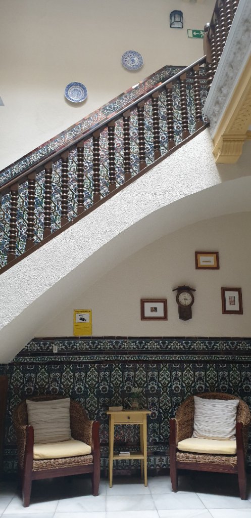 A central area showcases a curved staircase adorned with intricate Andalusian tiles. Two woven chairs are positioned beside a small wooden side table, while framed pictures and a clock are displayed on the wall, adding to the welcoming ambiance of the space.