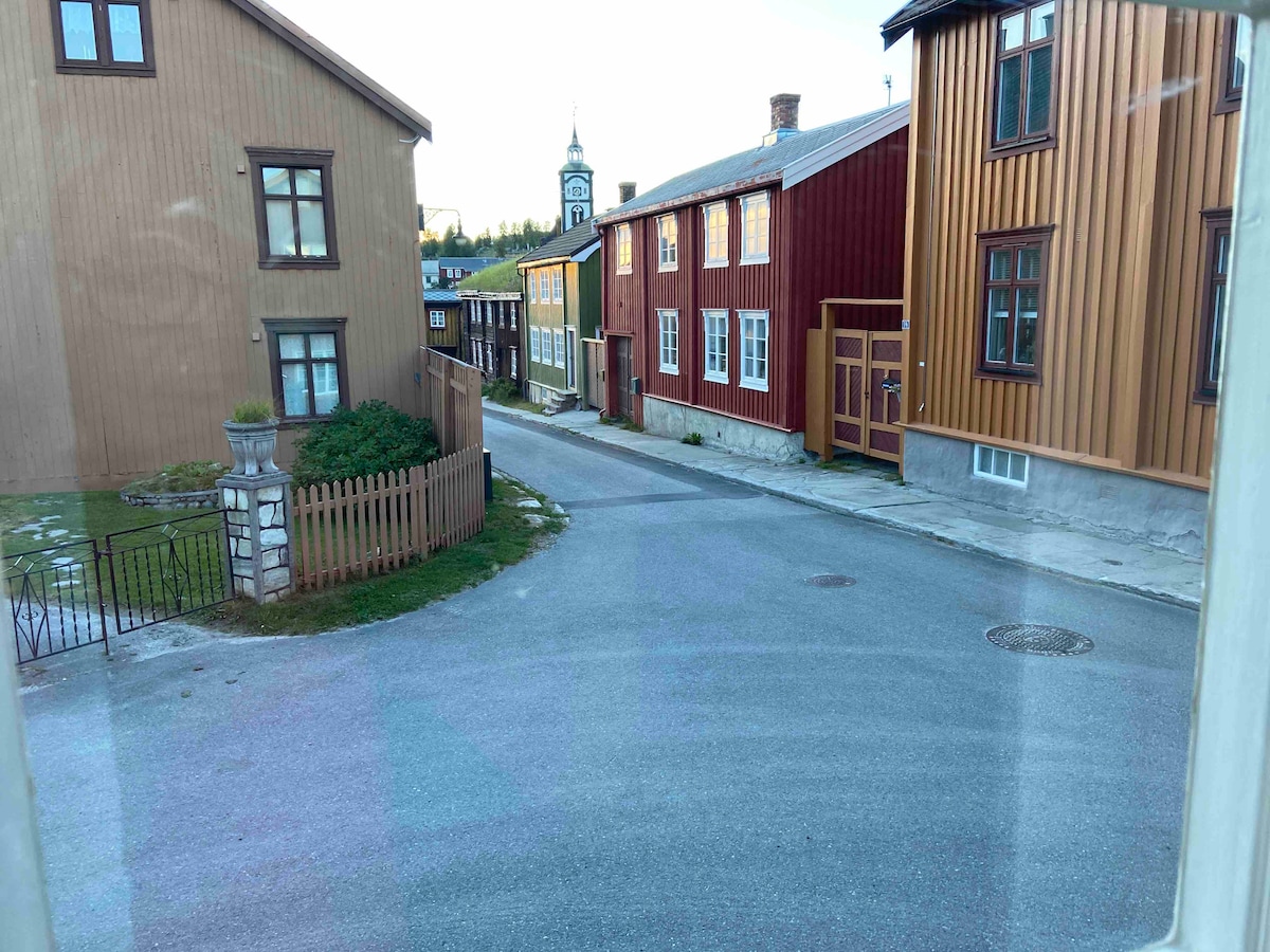 A view of a quiet street featuring a mix of wooden buildings in various colors including red and yellow. A clock tower is visible in the distance. The road curves gently, bordered by a low wooden fence and a grassy area with a stone feature.