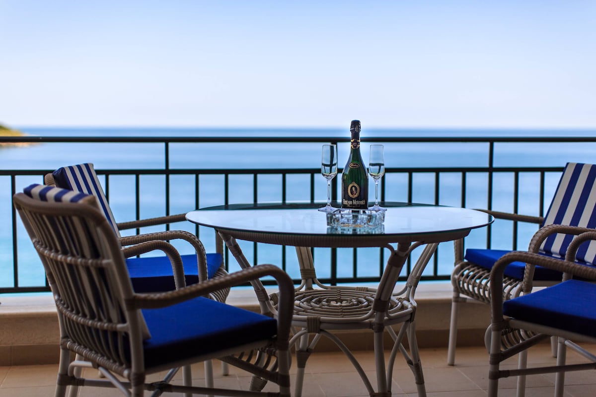 A terrace dining area features a round glass table surrounded by four cushioned chairs with blue upholstery. A bottle of champagne is placed on the table, accompanied by two glasses, while a serene view of the ocean extends into the distance.