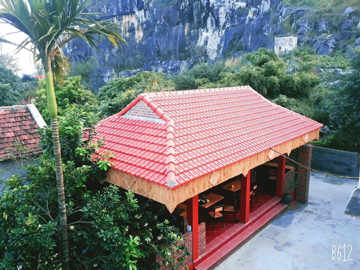 A traditional-style building with a vibrant red tiled roof is surrounded by lush greenery and natural rock formations. The structure features an open area with wooden tables and chairs, providing a communal dining space that blends seamlessly with the outdoor landscape.