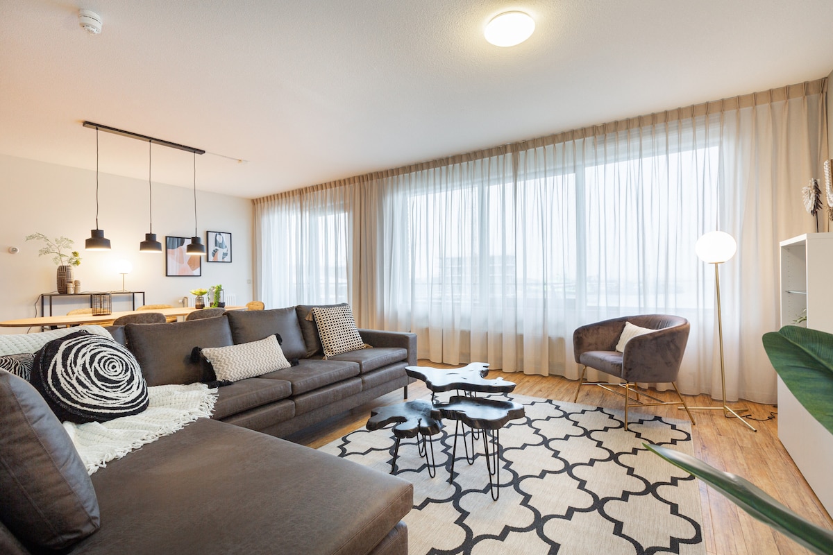 A spacious living area is illuminated by natural light filtering through sheer curtains. A dark sectional sofa is accompanied by a decorative throw and pillows, while modern side tables add a touch of elegance. A cozy armchair and a minimalist floor lamp enhance the inviting atmosphere.