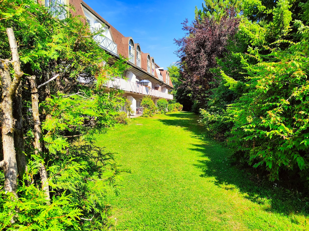 A well-maintained green lawn stretches in front of a two-story building, surrounded by dense foliage. The balconies of the building are visible on the upper level, providing outdoor spaces for relaxation among the greenery. A clear blue sky frames the scene.