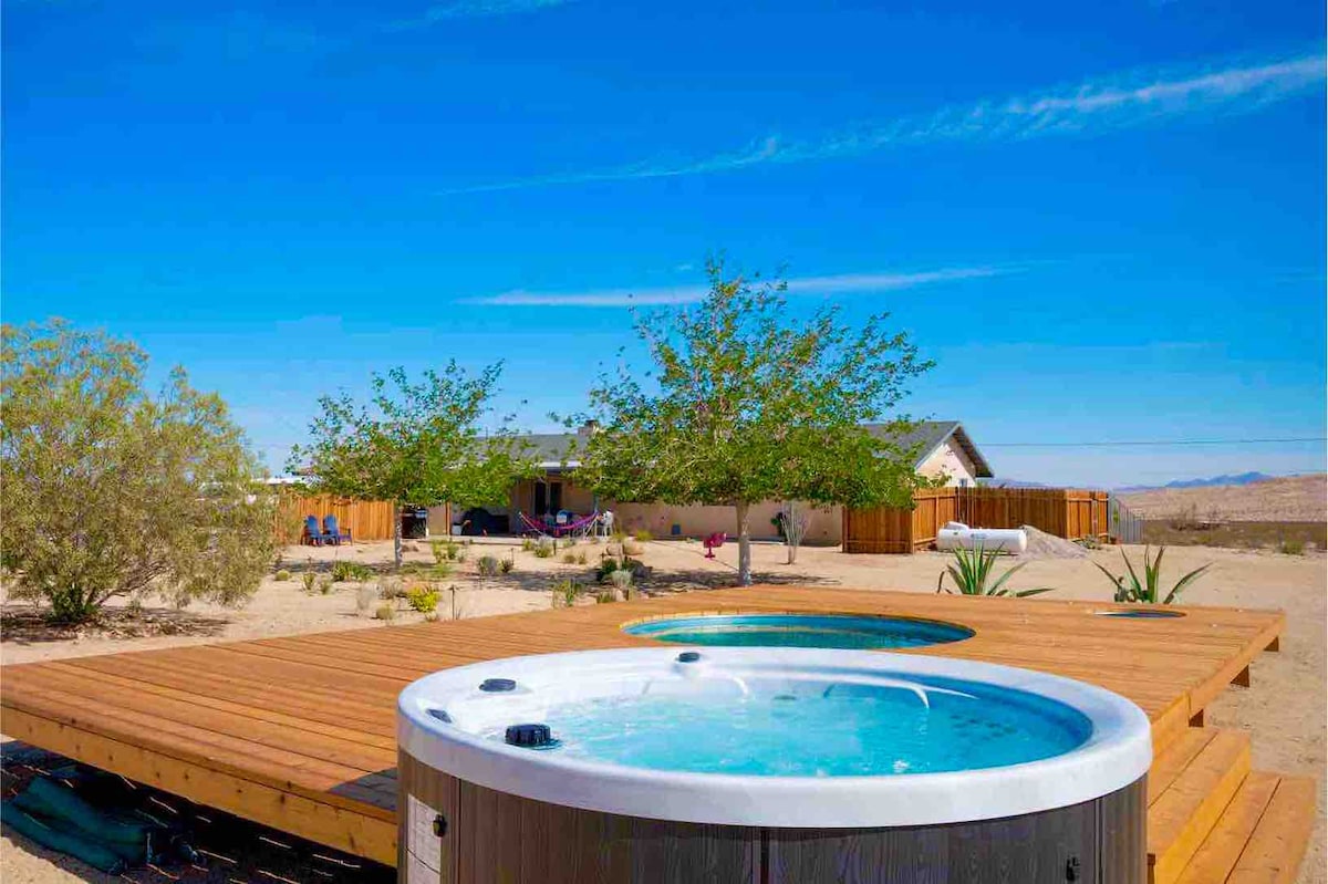 An elevated wooden deck features a stylish hot tub and a cowboy pool, surrounded by natural desert landscaping. Various shrubs and trees provide a sense of privacy while the clear blue sky creates a bright atmosphere, inviting relaxation and outdoor enjoyment.