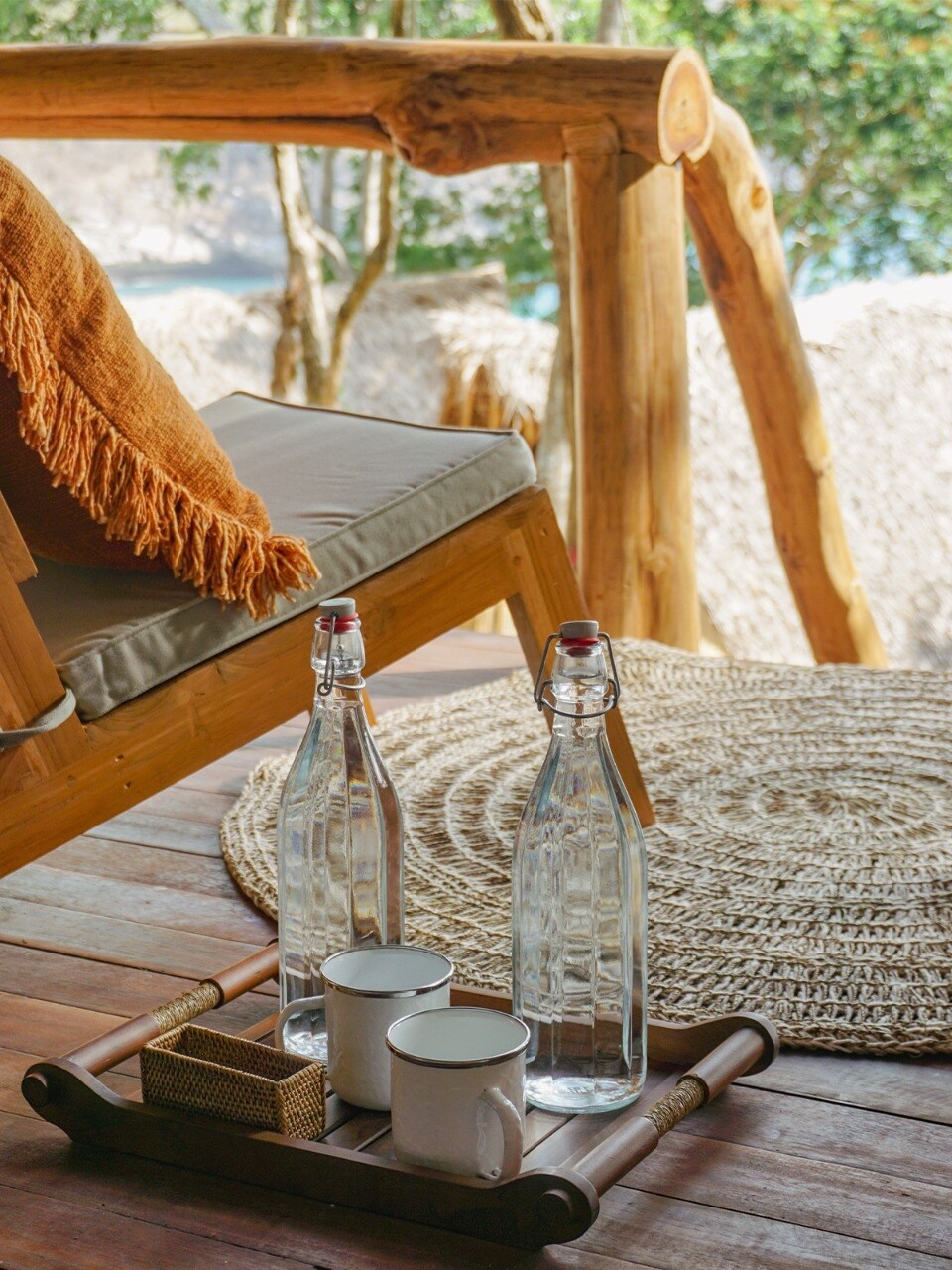 A wooden chair with a soft throw blanket is positioned next to a round woven rug. A wooden tray holds two glass bottles and two cups, creating a simple refreshment area. Natural light filters through nearby trees, enhancing the serene atmosphere.