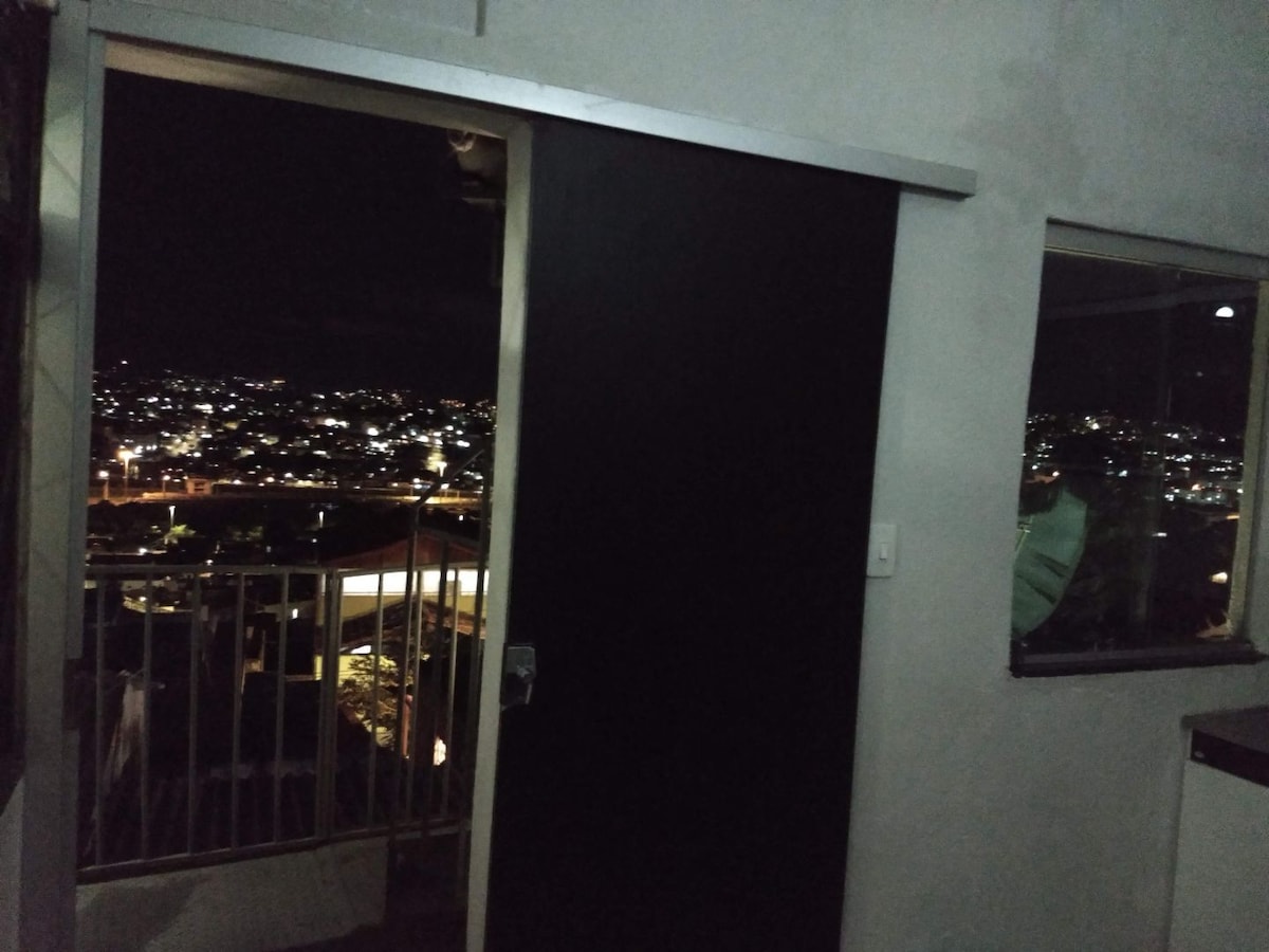 A dark sliding door is framed against a backdrop of city lights visible through a large window. The night view showcases a sprawling urban landscape, with twinkling lights reflecting off surfaces in the room. The walls are minimalist, enhancing the view outside.