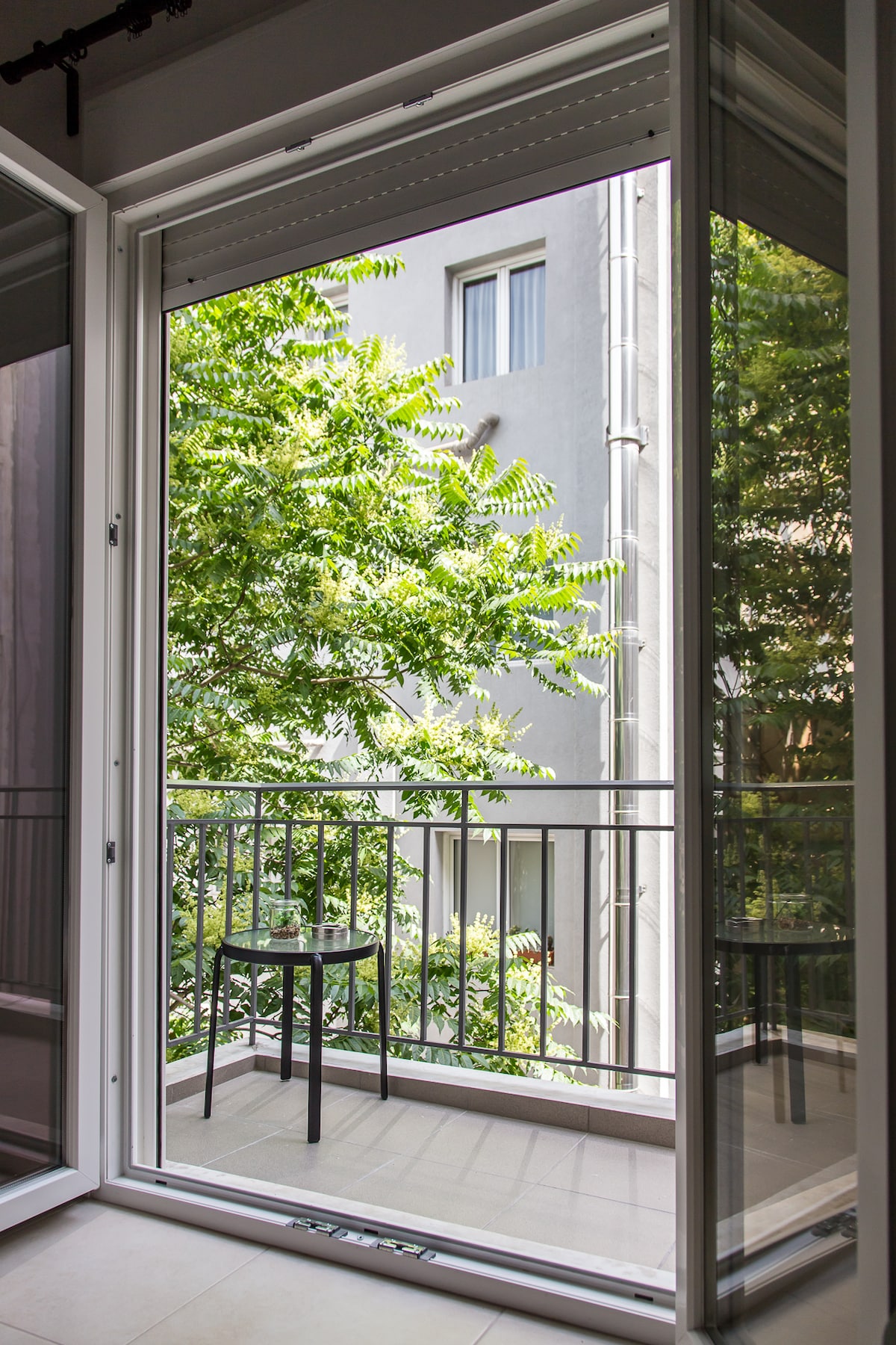 Large glass doors open to a small balcony, revealing a view of lush green foliage. A compact table with a single chair sits on the balcony, providing a serene outdoor space. The backdrop features a light-colored building, enhancing the natural light in the area.