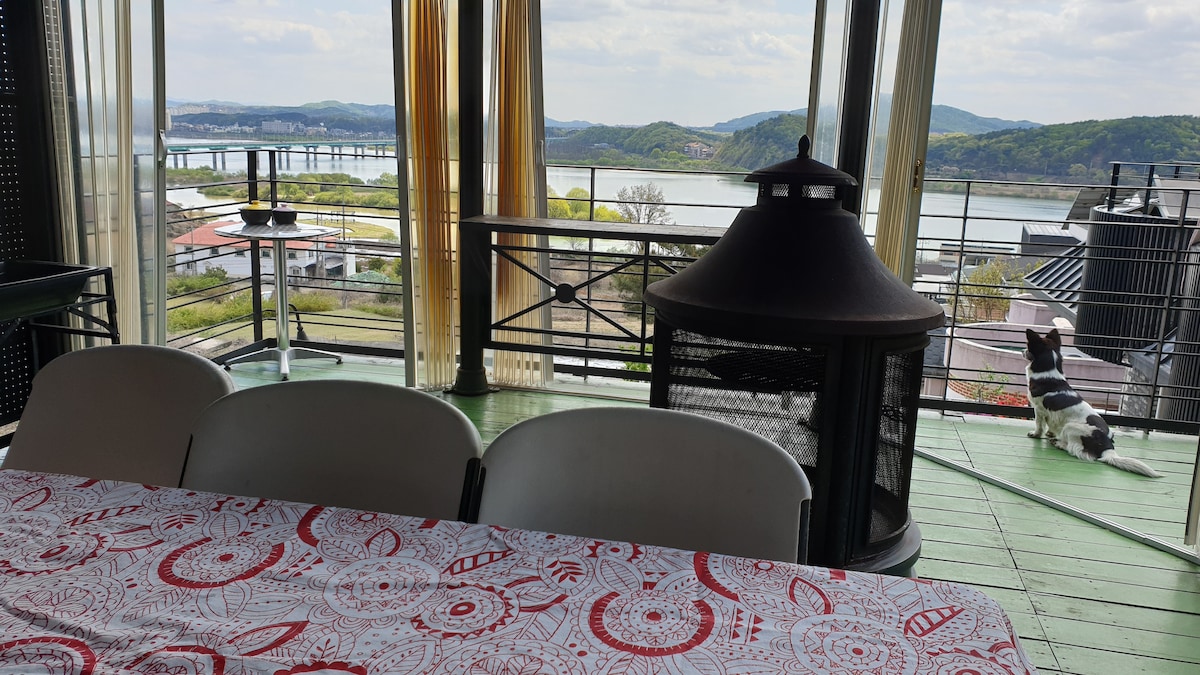 A spacious dining area features a table set against large glass doors, providing views of the tranquil river and surrounding hills. A modern fireplace adds a cozy touch to the space, while a dog can be seen relaxing nearby.