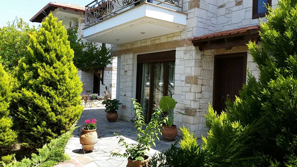 The exterior of the house is framed by lush greenery and potted plants. Stone walls and a tiled roof create a harmonious blend with the natural surroundings. Large windows are seen, allowing for ample natural light. A pathway leads to the entrance, surrounded by vibrant flowers.