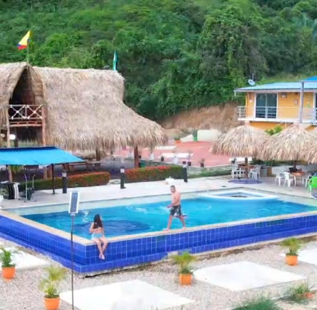 An expansive outdoor pool is surrounded by lush green hills. Cabana-style structures with thatched roofs provide shaded sitting areas. Several lounge chairs are positioned around the pool, offering a space for relaxation. A few guests can be seen enjoying the water, enhancing the inviting ambiance.