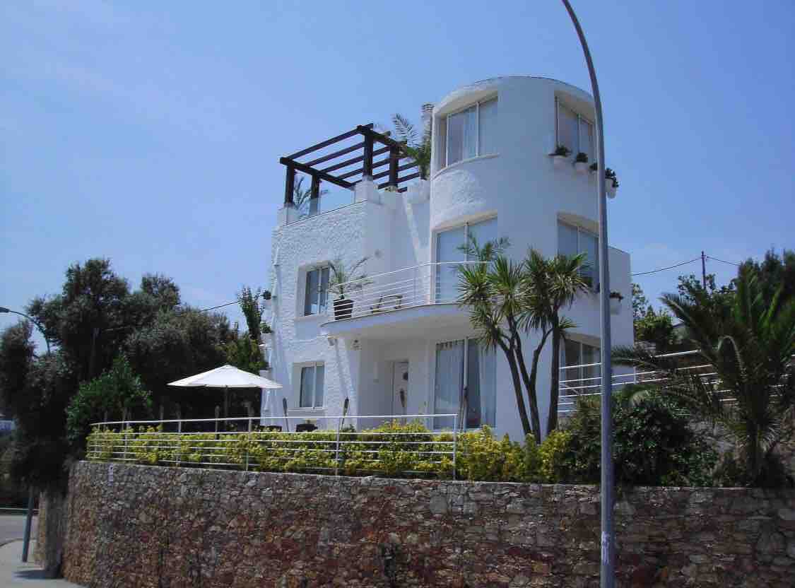 The villa features distinctive architecture with a white facade and curved corners. A rooftop terrace is visible with wooden beams and plants. Lush greenery and palm trees frame the property, while a light stone wall adds contrast to the inviting outdoor space.