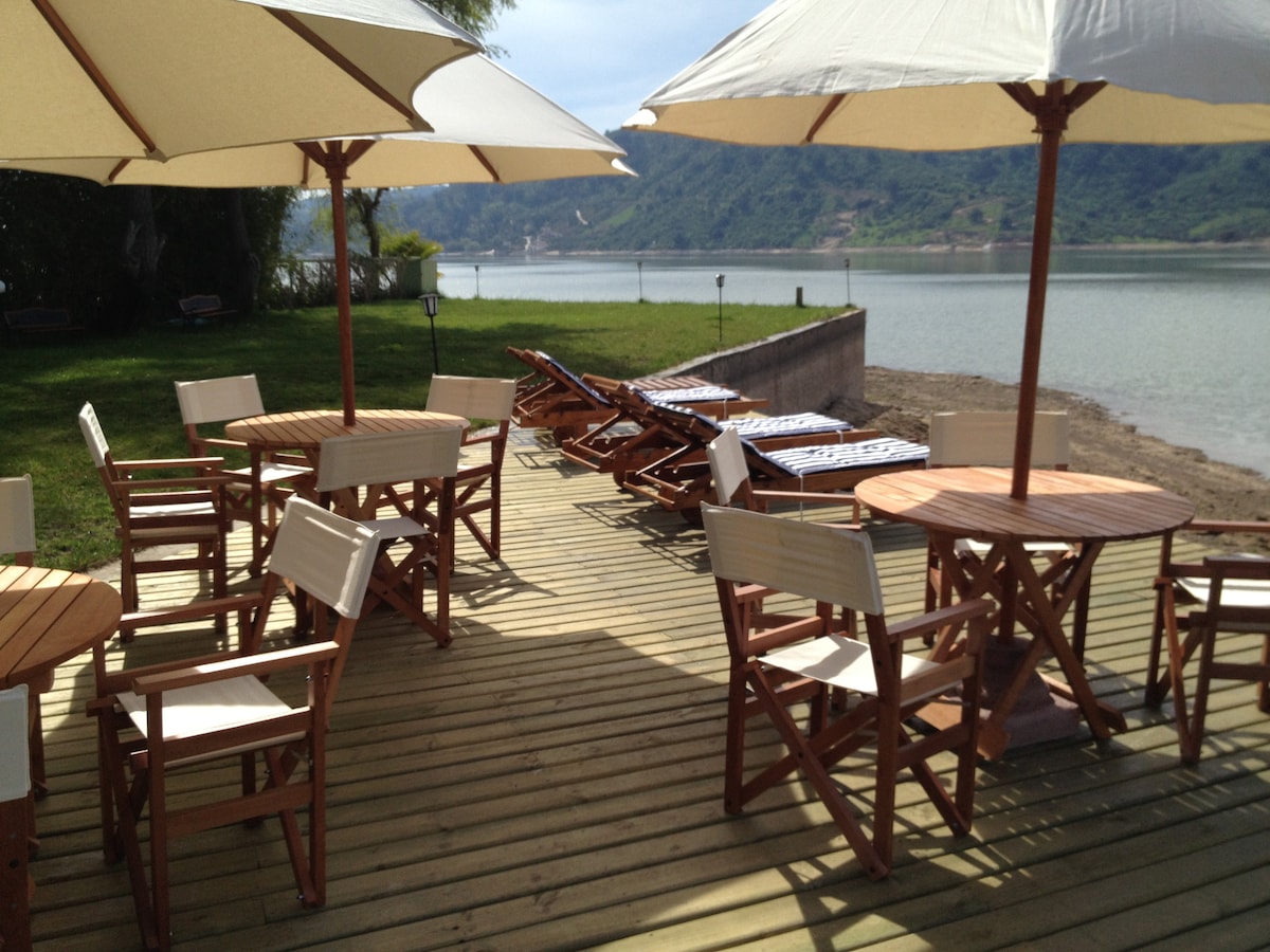 A wooden deck beside the lake features several tables and chairs arranged under large umbrellas. Sun loungers are positioned nearby, providing a comfortable spot for relaxation while enjoying the scenic view of the water and surrounding hills.