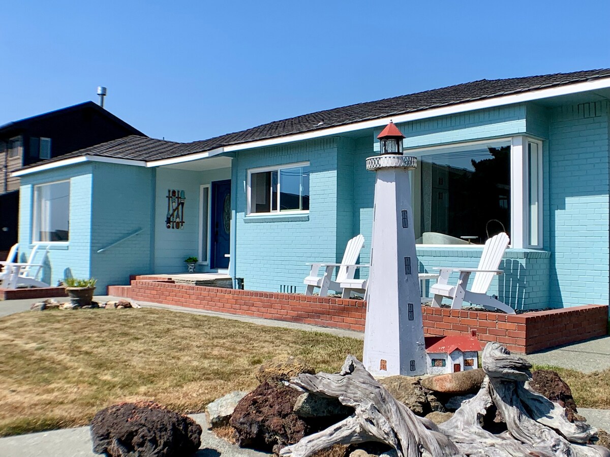The exterior of the beach house showcases a light blue façade complemented by large picture windows. A decorative lighthouse sculpture is positioned in the foreground, surrounded by driftwood and a small garden. Adirondack chairs are arranged on a patio, inviting relaxation.