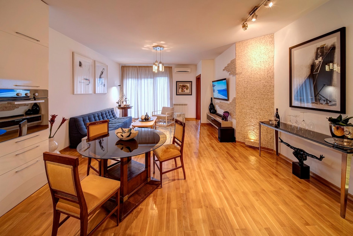 The living area features a stylish sofa and a dining table surrounded by wooden chairs. Natural light flows through large windows, illuminating the wooden flooring. A flat-screen television is mounted on the wall, and artwork decorates the space, adding modern touches.