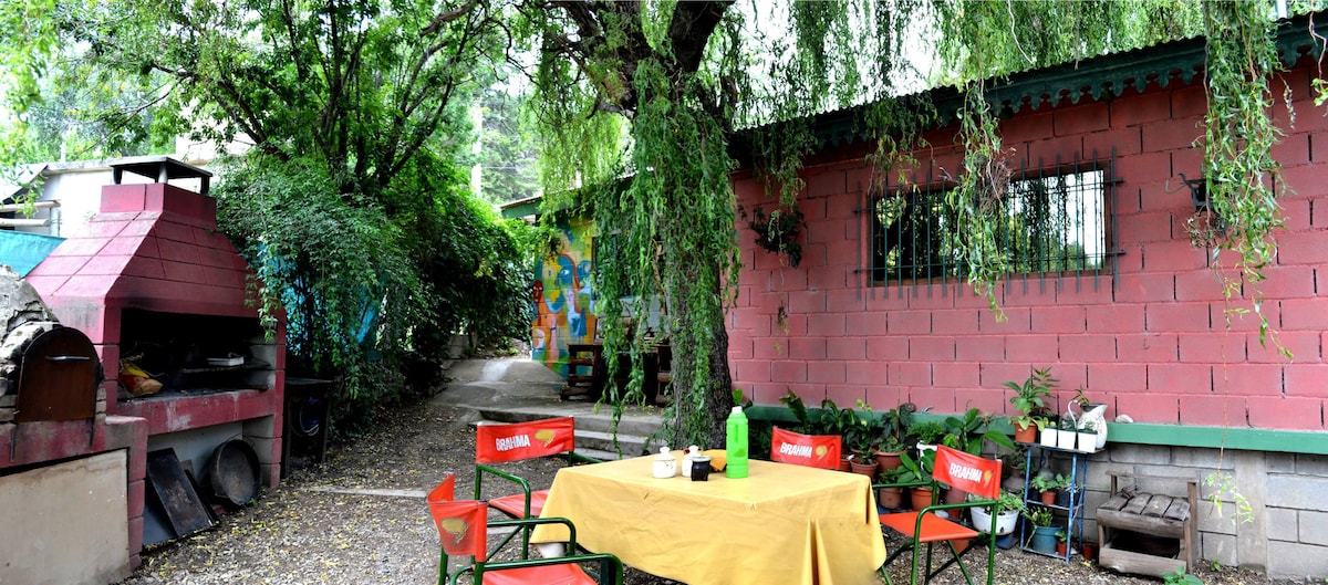 An outdoor patio area is surrounded by lush greenery, featuring a table with a yellow tablecloth set for dining. Five folding chairs are positioned around the table. A brick grill and a vibrant mural are visible in the background, adding color to the harmonious setting.