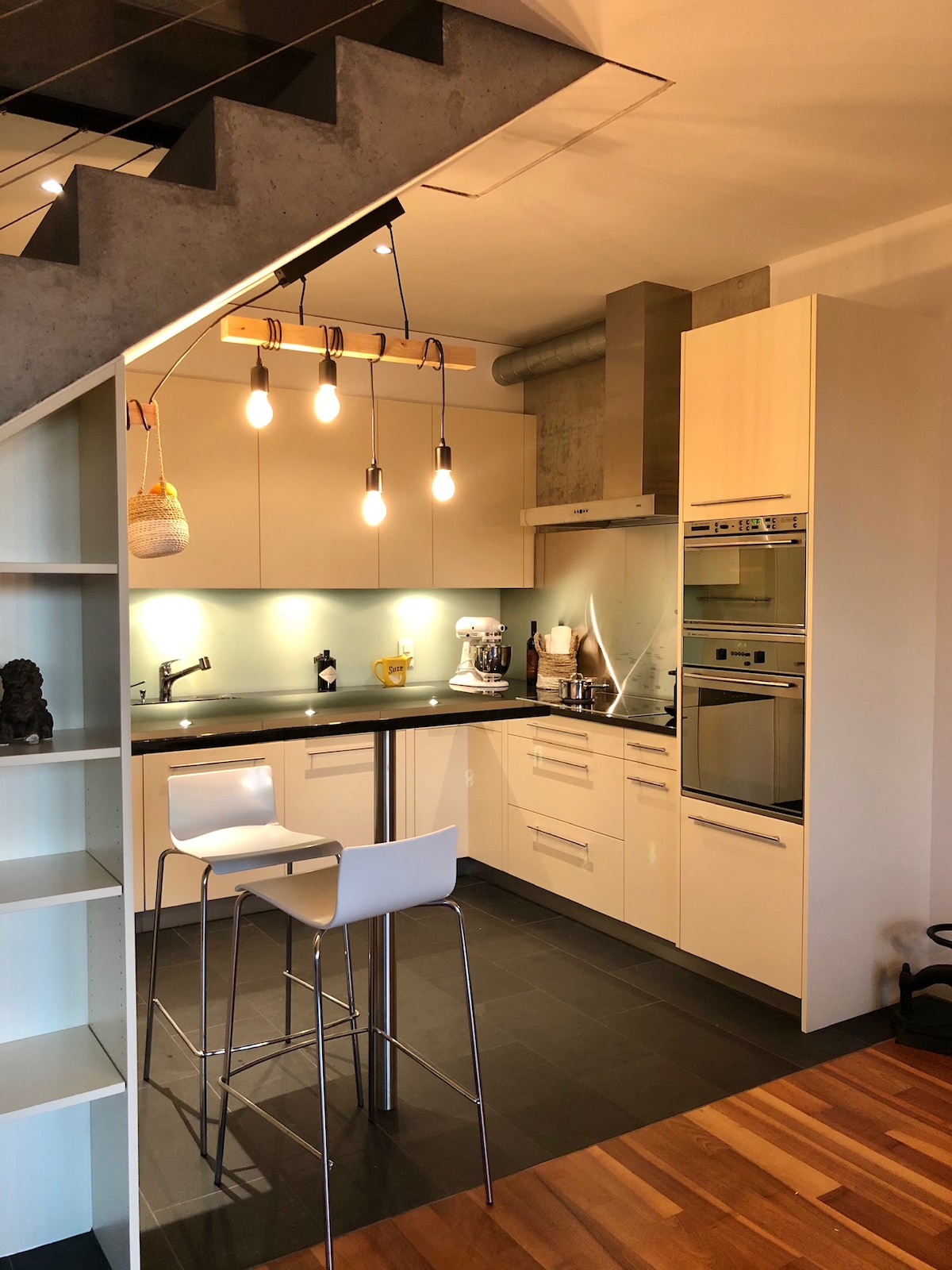 A modern kitchen area is highlighted by sleek cabinetry and stainless-steel appliances, creating a functional cooking space. An island with high stools provides a spot for casual dining. Warm wood flooring contrasts with the black tile, enhancing the contemporary design.