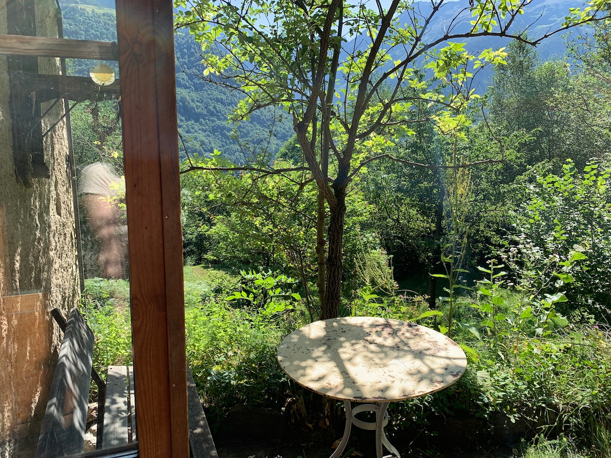 A small outdoor seating area is visible, featuring a round table surrounded by lush greenery. A tree provides shade, and rolling hills can be seen in the background, creating a calm, natural setting.