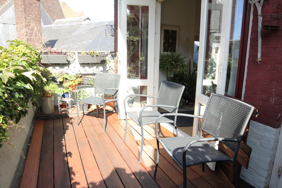 The balcony features a wooden deck with several black metal chairs arranged for relaxation. Lush green plants are positioned around the space, providing a touch of nature. Large glass doors lead into the living area, creating a bright and open ambiance.