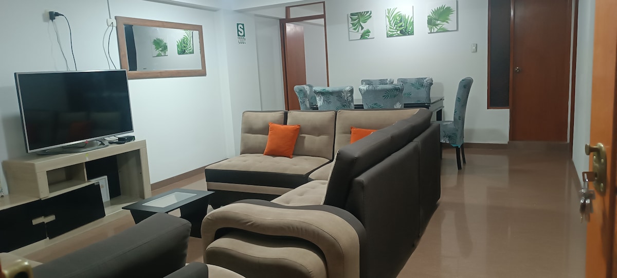 A spacious living area is showcased, featuring a comfortable seating arrangement with a sectional sofa and armchairs. A television is mounted on the wall opposite a glass coffee table. A dining table with upholstered chairs is positioned in the background, complemented by framed greenery artwork.