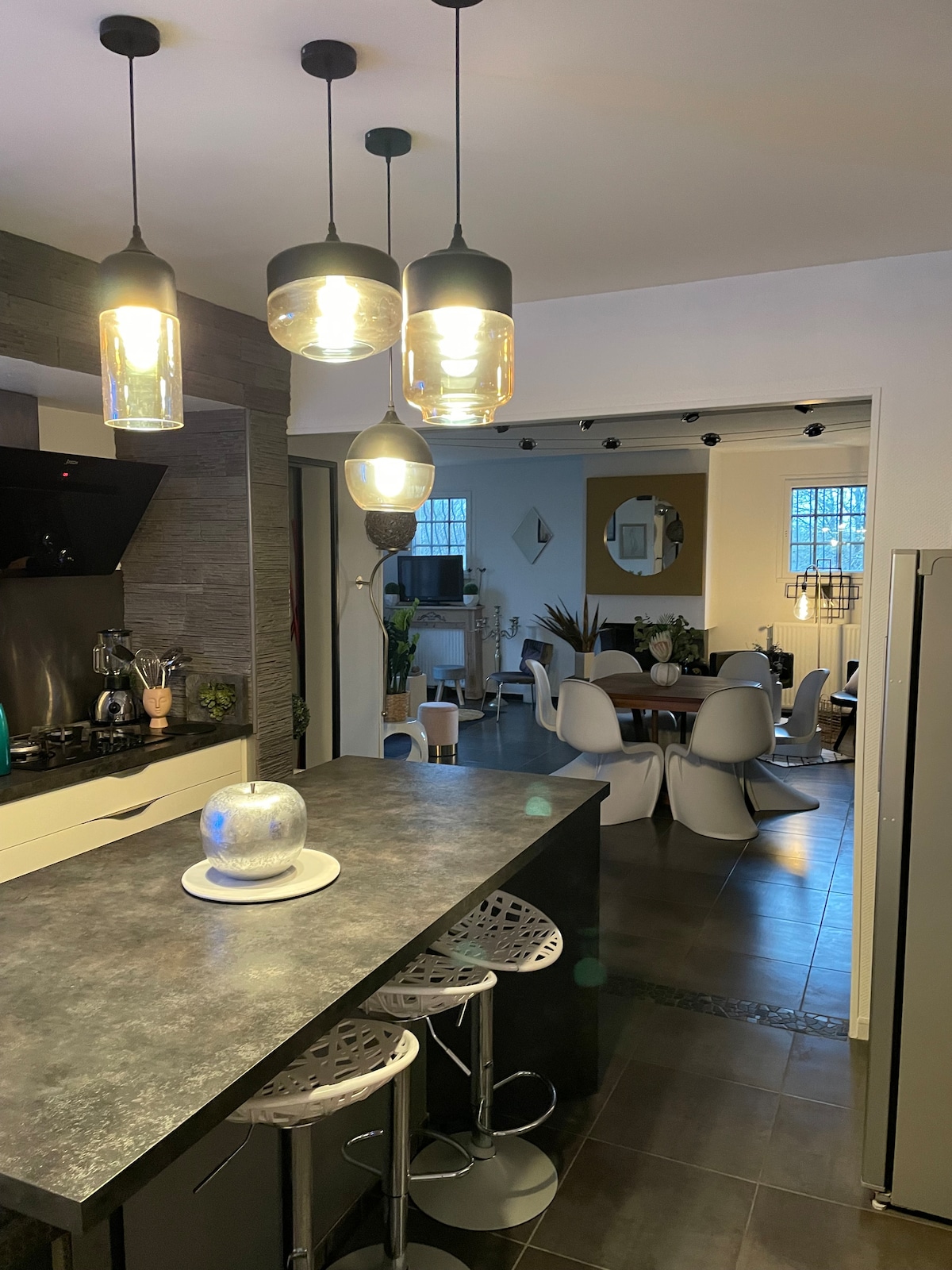 A modern kitchen area showcases a dark countertop with bar stools. Three pendant lights hang above the countertop, illuminating the space. The open layout leads to a dining area, where sleek chairs surround a round table, and natural light filters through a window.