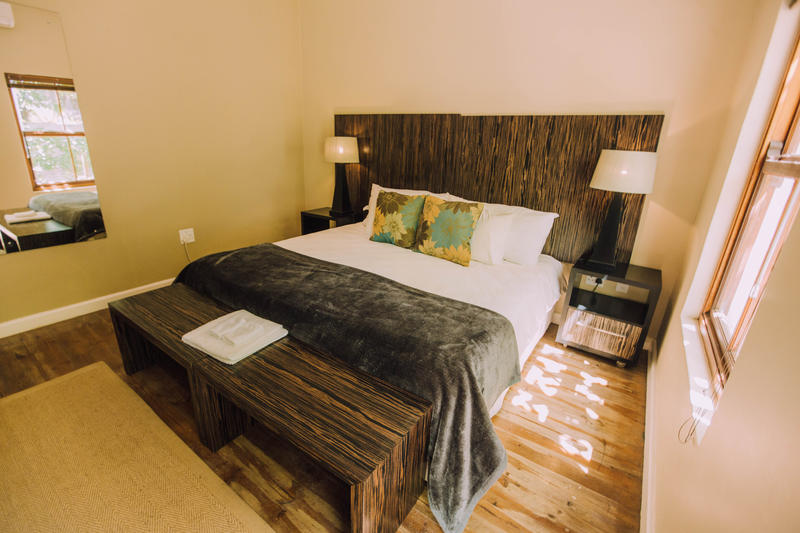 A tastefully furnished bedroom features a spacious bed with decorative pillows against a wood-paneled headboard. Soft lighting is provided by bedside lamps. A wooden bench is positioned at the foot of the bed, and natural light enters through windows with a view of the room's warm, neutral walls.
