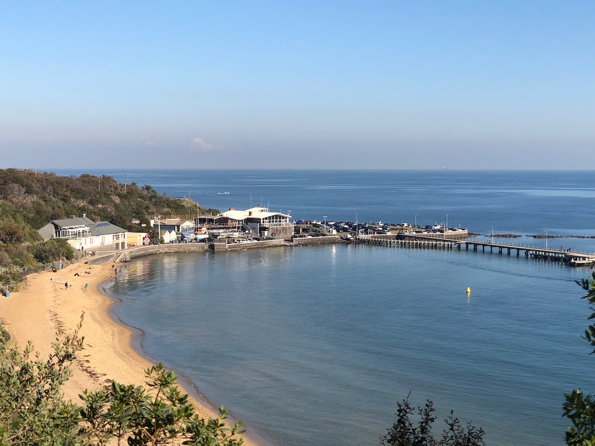A serene beach scene captures the gentle curve of the shoreline, bordered by golden sand. Calm waters reflect the clear blue sky while a pier extends into the bay. Various buildings are visible near the water's edge, contributing to the tranquil atmosphere.