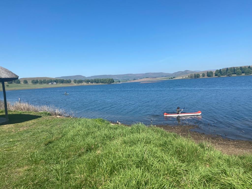 A serene view of the Springrove dam is presented, with clear blue waters and a gentle breeze across the surface. A canoe is moored near the grassy shore, while a distant figure paddles on the water. The backdrop features rolling hills under a clear sky.