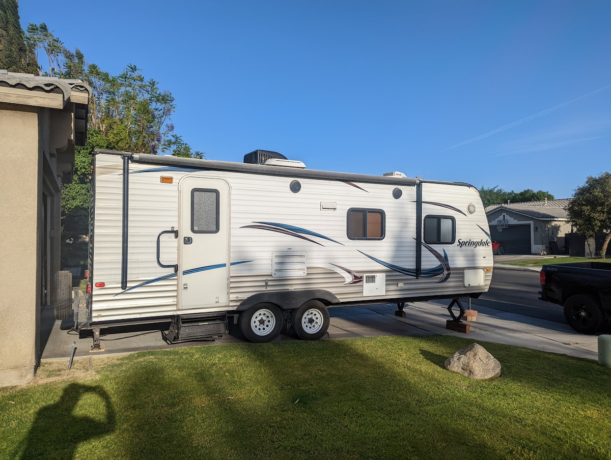 The RV is parked on a grassy area adjacent to a house, showcasing its exterior design with sleek lines and a light color scheme. A nearby driveway is visible, providing access for vehicles. The surrounding neighborhood includes trees and homes, indicating a quiet environment.