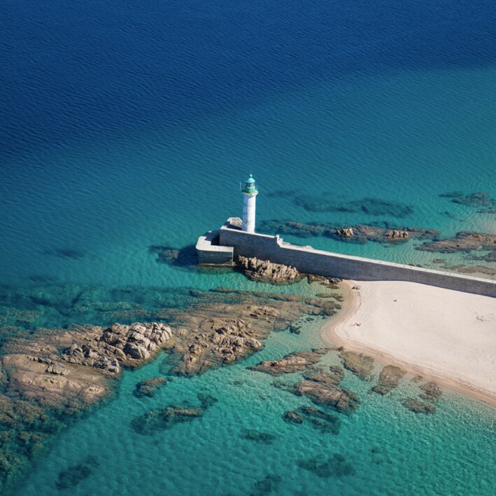 The image captures an aerial view of a coastal scene featuring a lighthouse standing on a stone pier, surrounded by clear turquoise waters. Rocky formations are visible beneath the surface, and a sandy beach is located at the water's edge.