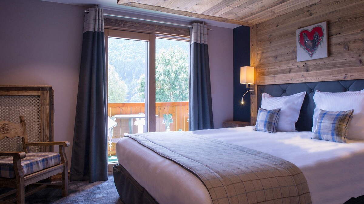 A cozy double room features a spacious bed with crisp white linens and decorative throw pillows. Natural light floods the space through sliding glass doors that open to a sun-exposed balcony with scenic mountain views. A wooden accent wall and rustic furnishings enhance the room's charm.