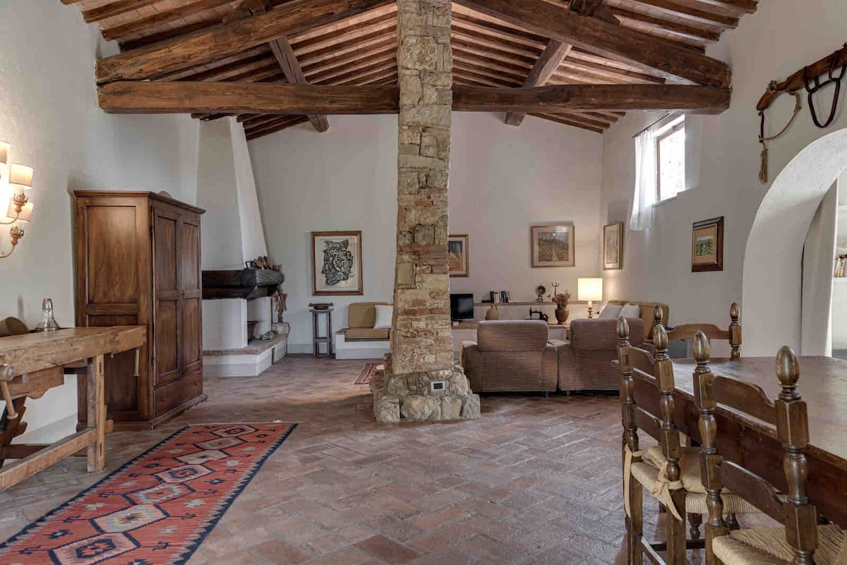 A spacious living area features rustic wooden beams and a stone pillar. A cozy lounge corner includes comfortable seating and a television, while a dining table with wooden chairs invites shared meals. The space is adorned with various artworks and framed pictures.