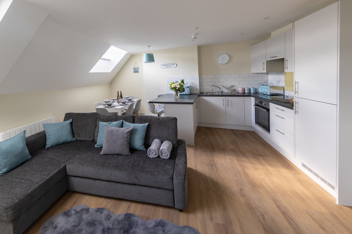 A modern open-plan living space features a corner sofa with plush cushions, surrounded by natural light from a skylight. A sleek kitchen area is visible, equipped with integrated appliances. A dining table for four is set nearby, creating an inviting space for meals.
