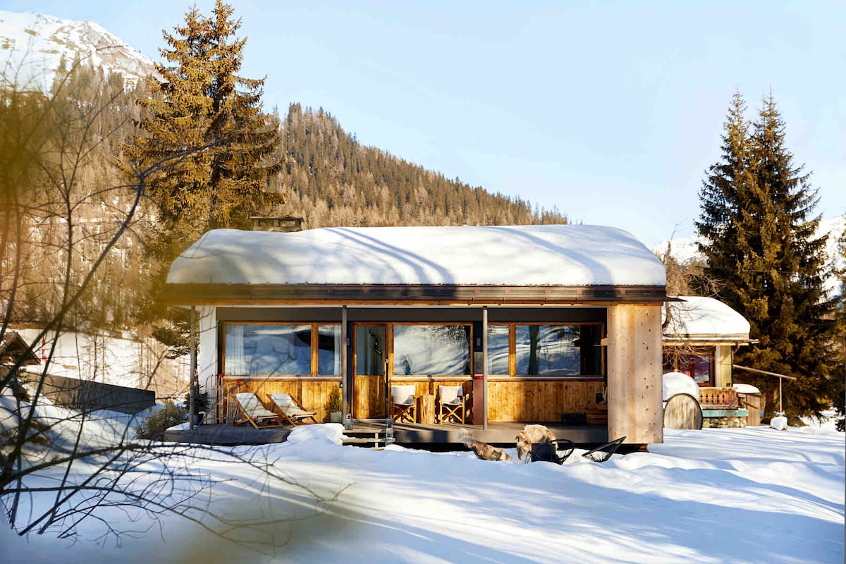 A cozy chalet is surrounded by snow-covered terrain, featuring large windows that invite natural light. The sloping roof is blanketed with fresh snow, while wooden deck chairs provide a welcoming space to enjoy the serene alpine landscape.