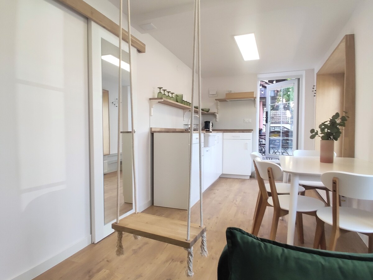 An open-concept living area features a wooden swing suspended from the ceiling, offering a playful touch. A light wooden dining table with four chairs is positioned near a bright window, while a modern kitchen is visible in the background, equipped with sleek white cabinetry.