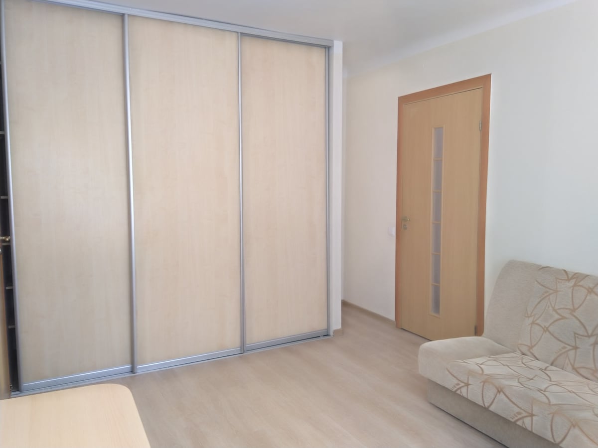 A light and airy interior features a spacious sliding door wardrobe made of light wood. A simple sofa with a patterned fabric sits against a wall, providing a comfortable seating area. The room is brightened by natural light, enhancing the overall ambiance.